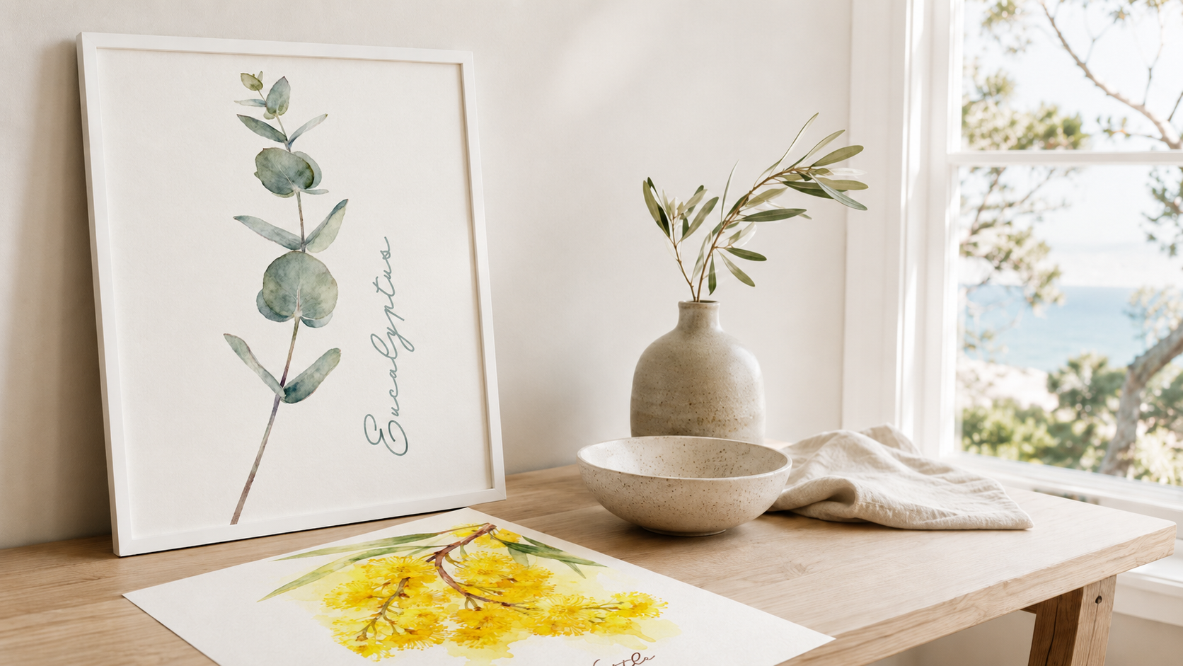 A framed eucalyptus print leaning on a light timber styling table beside a bright coastal window, with a wattle print, ceramic textures, olive foliage, linen details, and soft natural light.